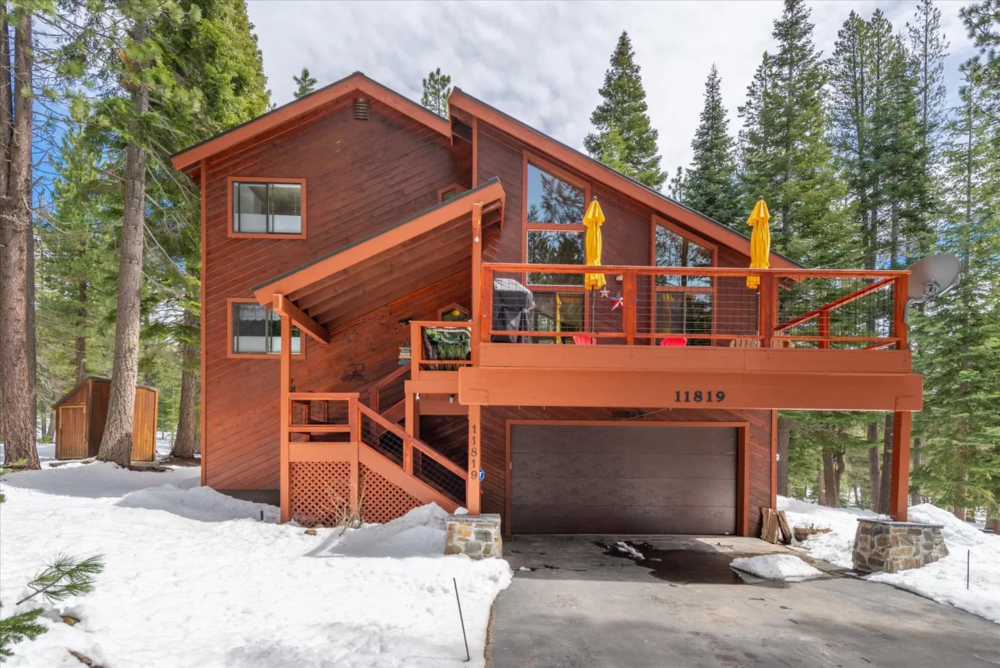A two-story brown house with a deck and garage, surrounded by snow and tall green trees.