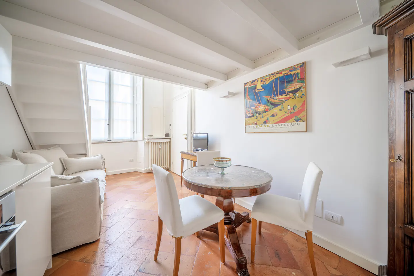 Bright living area with terracotta floors, a round dining table, white chairs, a sofa, and a colorful landscape painting.