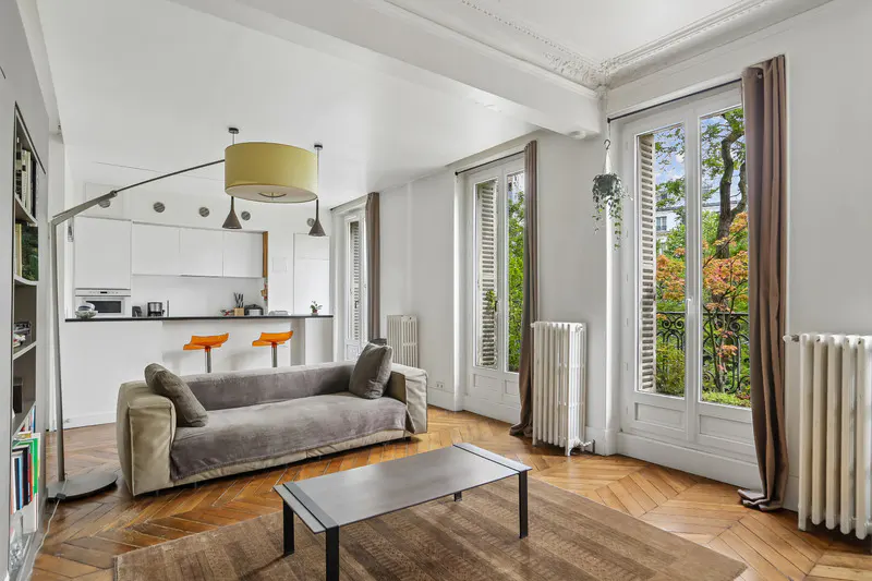 Bright, airy living room with parquet floors, a gray sofa, and a modern kitchen in the background. Large windows offer a view of lush greenery.
