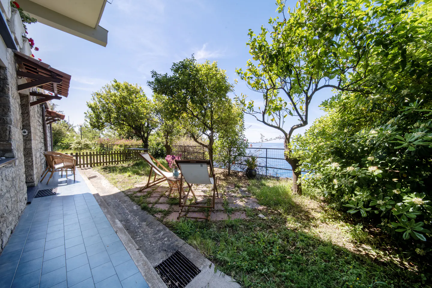 Patio with blue tile, stone wall, and outdoor seating. Trees and a glimpse of the ocean are in the background.