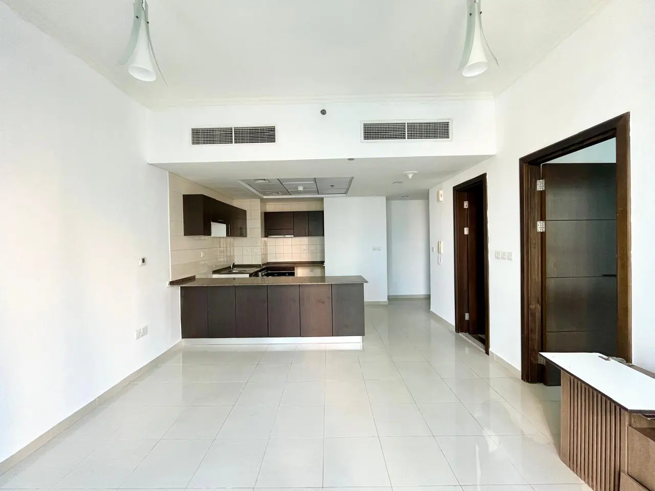Bright, empty apartment with white walls and tile floors. A dark brown kitchen with a breakfast bar is visible. Two dark wood doors are on the right.