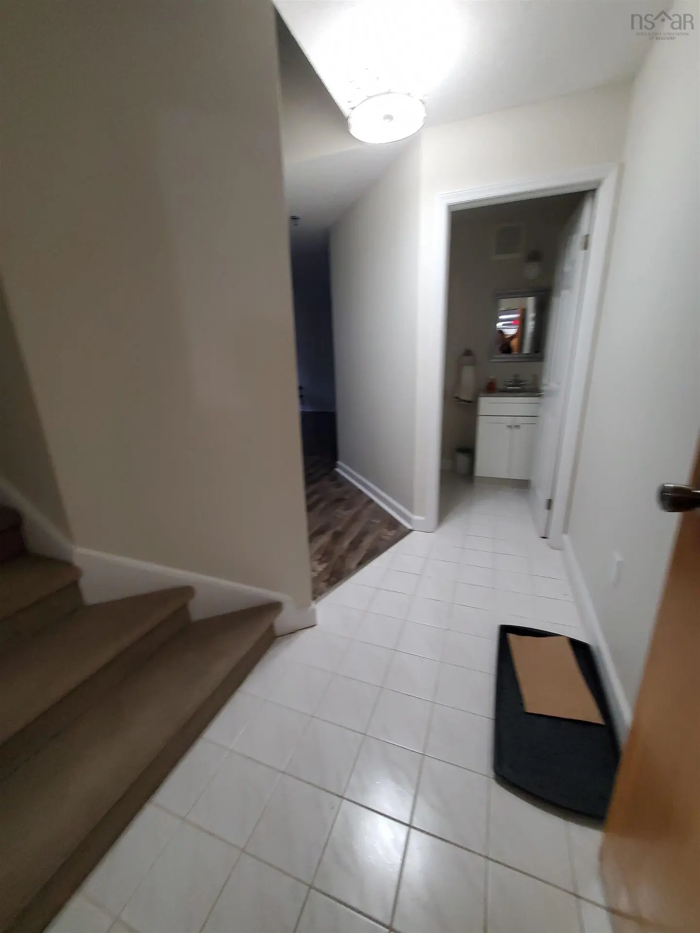 Entryway with white tile floor, stairs on the left, and a bathroom visible through a doorway. A black mat is on the floor.
