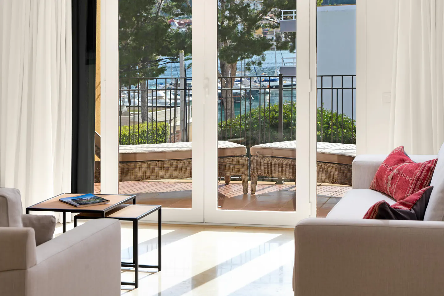 Bright living room with white sofa, red pillows, and nesting tables. Balcony with wicker furniture overlooks a harbor with boats.