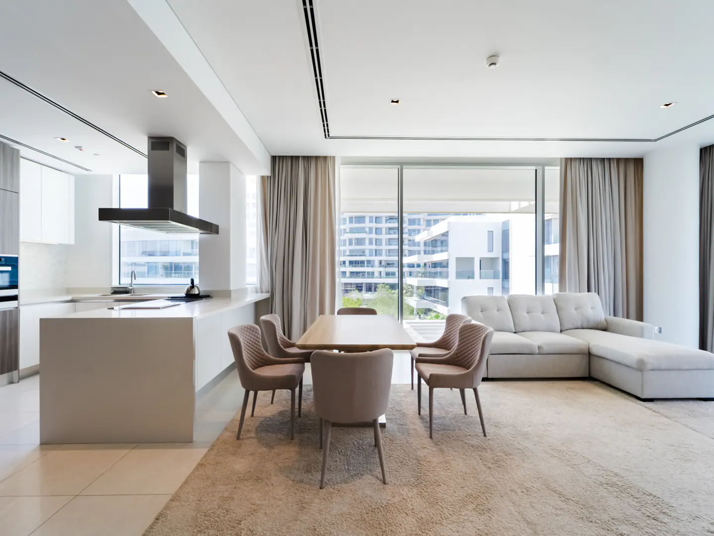 Modern open-plan living area with a white kitchen island, dining table with four chairs, and a light gray sectional sofa.