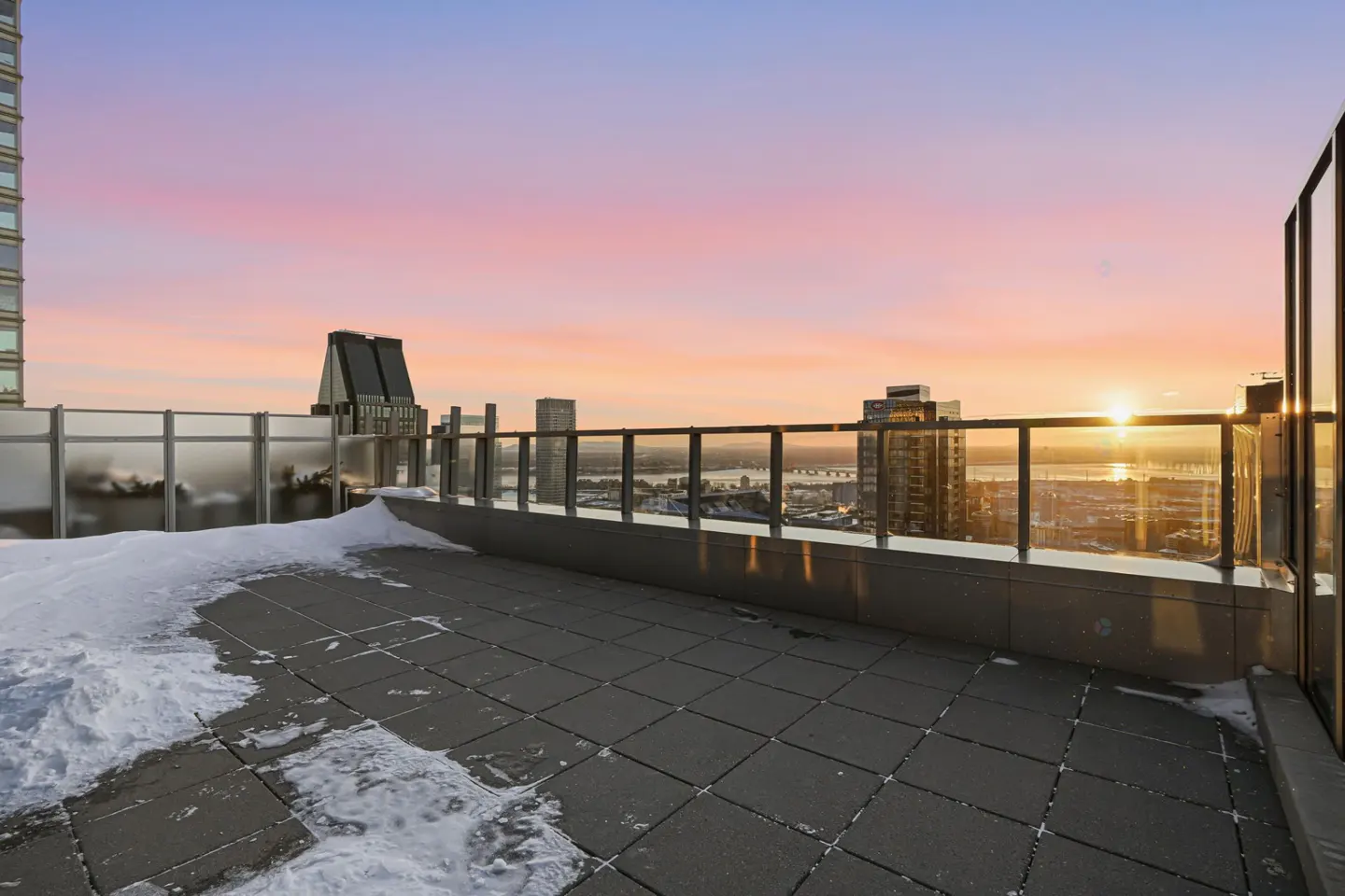 Rooftop patio with glass railing overlooks a city skyline at sunset. Snow is visible on the patio floor.
