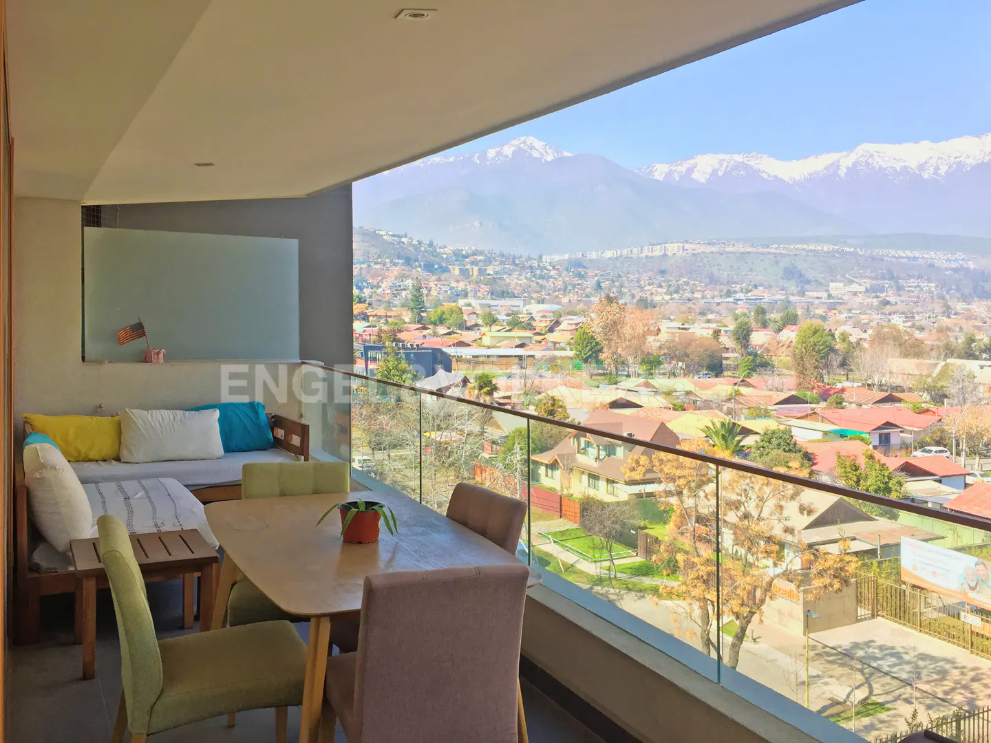 Balcony with table, chairs, and cushioned seating overlooks a city with snow-capped mountains in the distance on a sunny day.