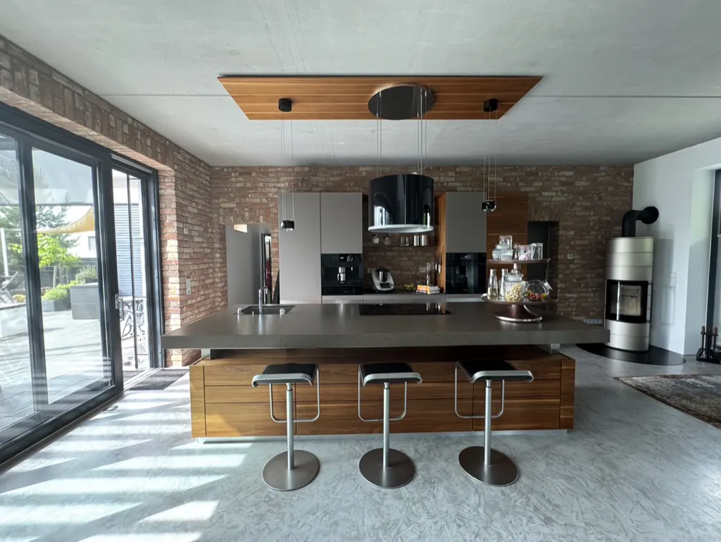 Modern kitchen with a large island, three bar stools, brick walls, and a cylindrical fireplace in an open-plan space.