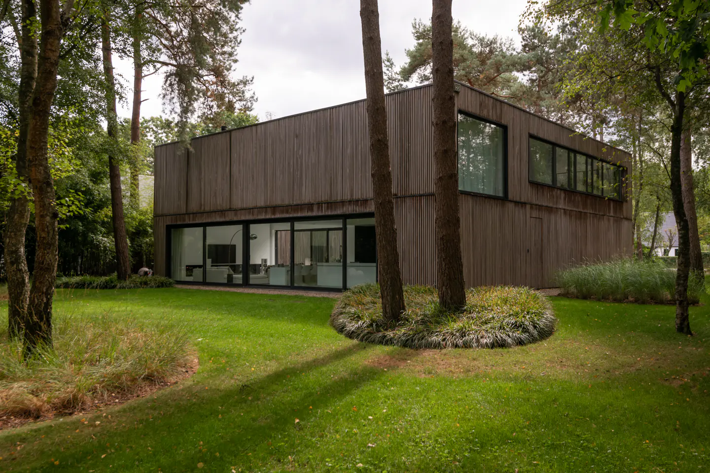 Modern two-story home with vertical wood siding, large glass windows, and a lush green lawn surrounded by tall pine trees.