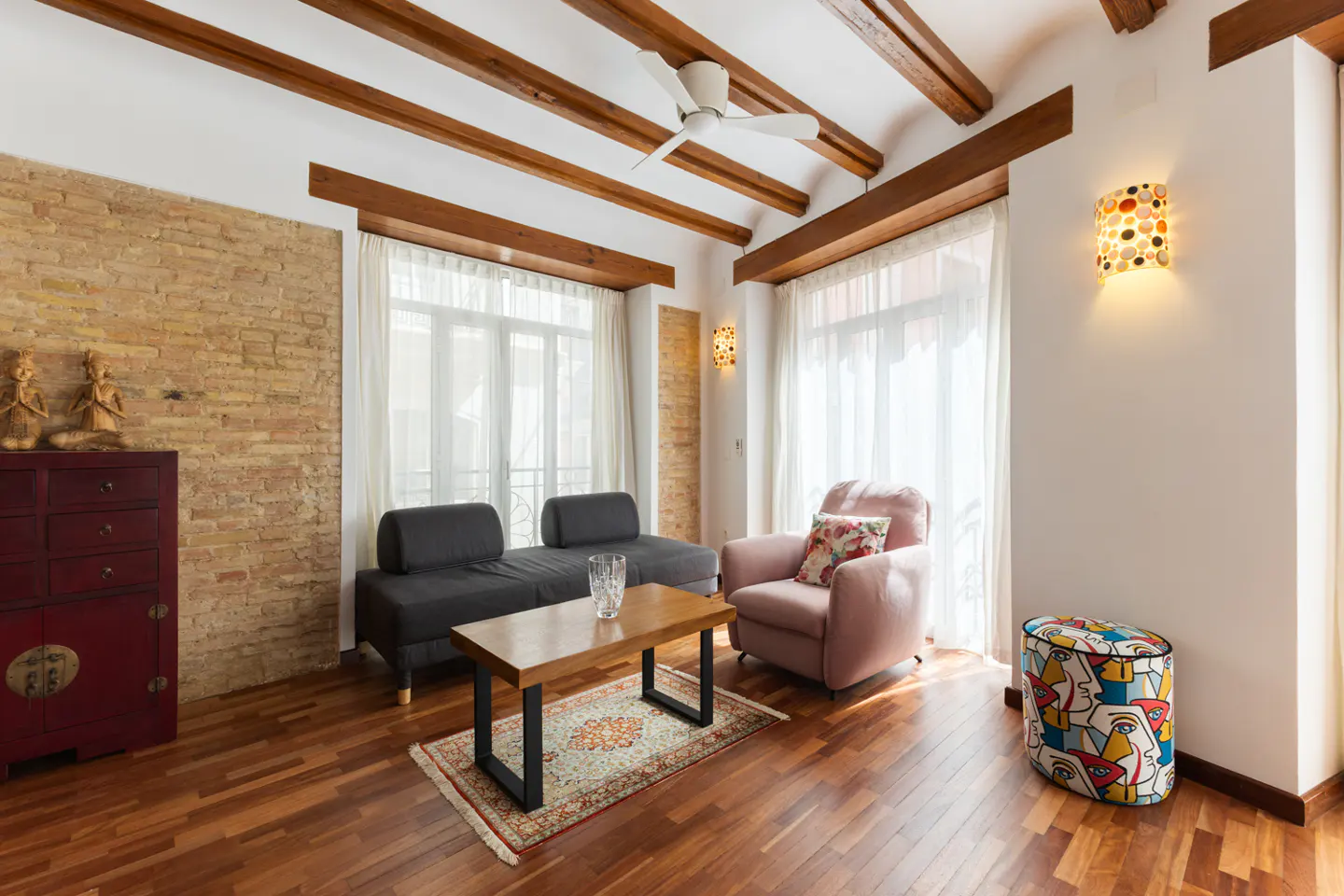 Living room with wood floors, exposed beams, and brick wall. A gray sofa, pink armchair, and patterned ottoman furnish the space.