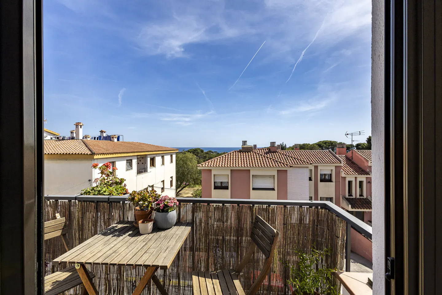 Balcony view with a wooden table, chairs, and potted flowers. Buildings with red tile roofs and a blue sky are in the background.