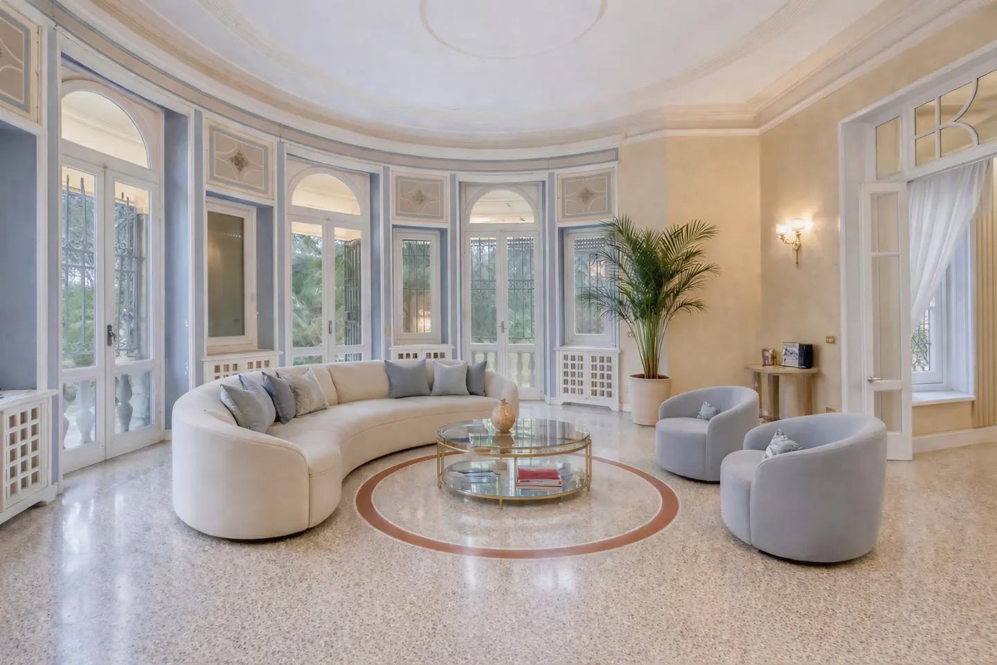 Bright, airy living room with curved cream sofa, blue chairs, and glass table. Large windows offer outdoor views. Terrazzo flooring.