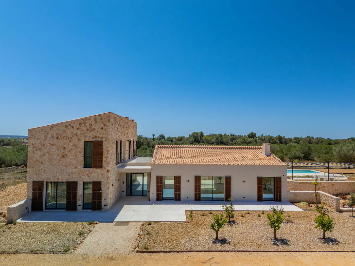 Modern stone and white villa with a tiled roof, large windows, and a swimming pool in a sunny, rural landscape.