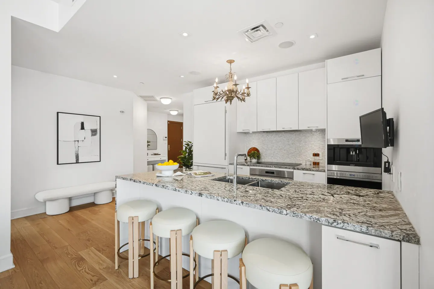 Bright kitchen with white cabinets, granite countertops, and four white leather bar stools. A gold chandelier hangs above the island. Hardwood floors and white walls.