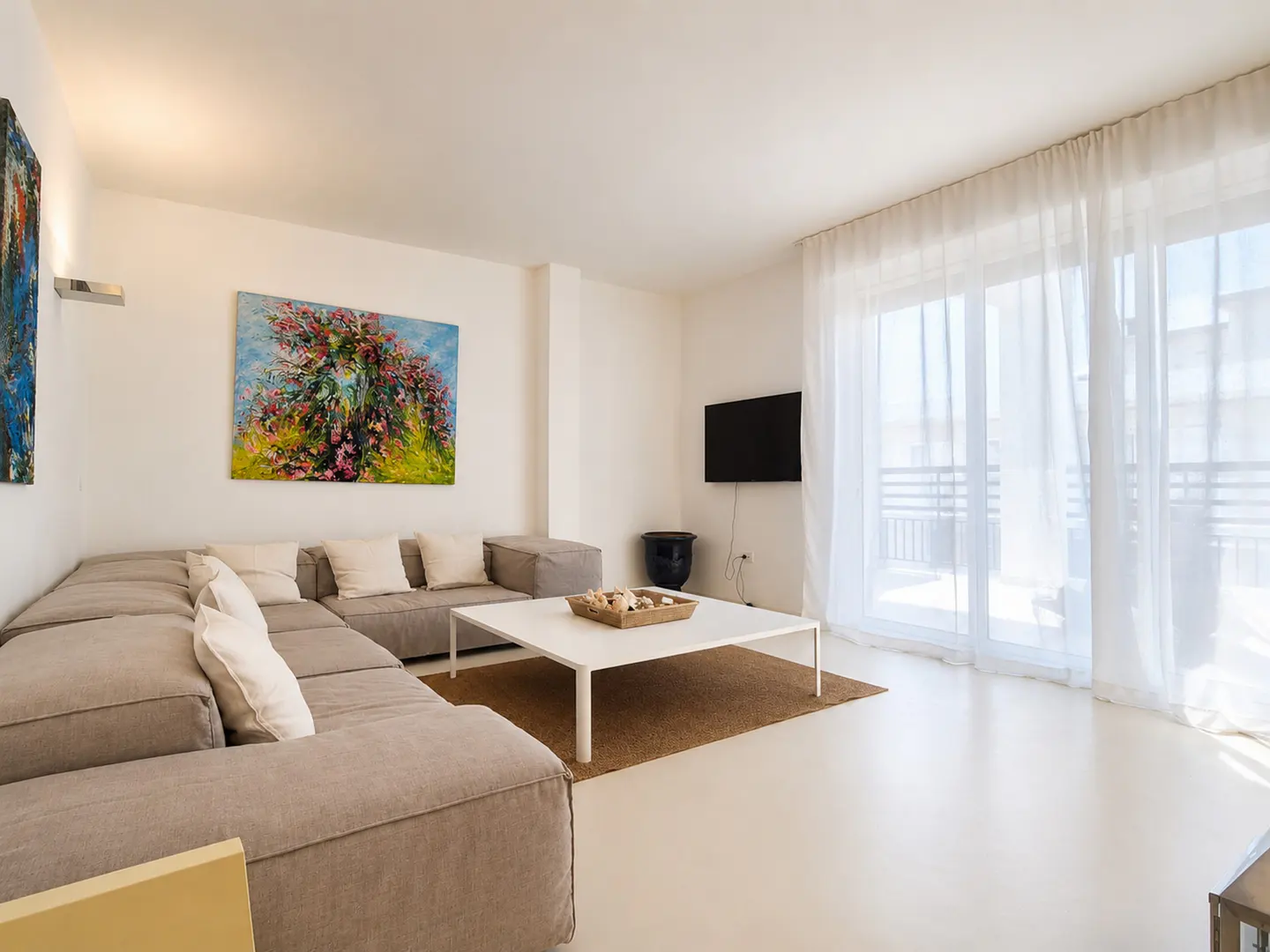 Bright living room with a large gray sectional sofa, white pillows, a white coffee table, and a colorful painting on the wall. Balcony with sheer white curtains.