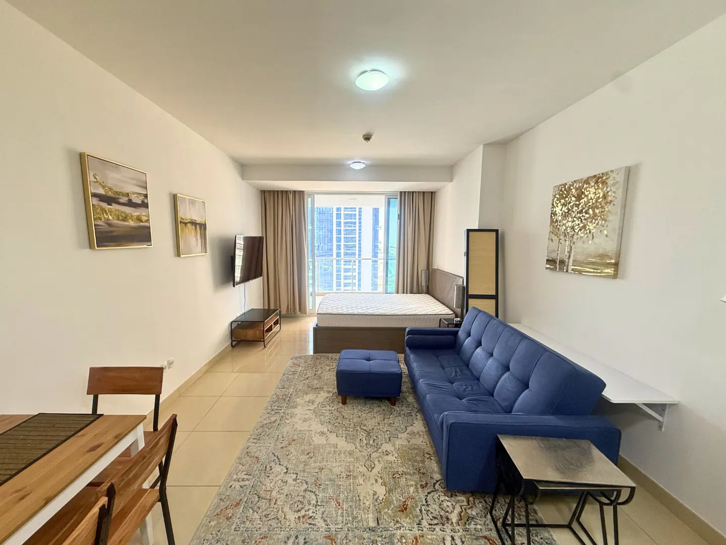 Bright studio apartment with a blue sofa, bed, and city view from the window. Art hangs on the white walls.