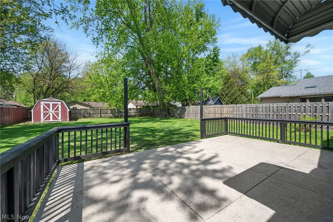 A backyard with a concrete patio, black railings, green grass, trees, and a red shed with white trim.