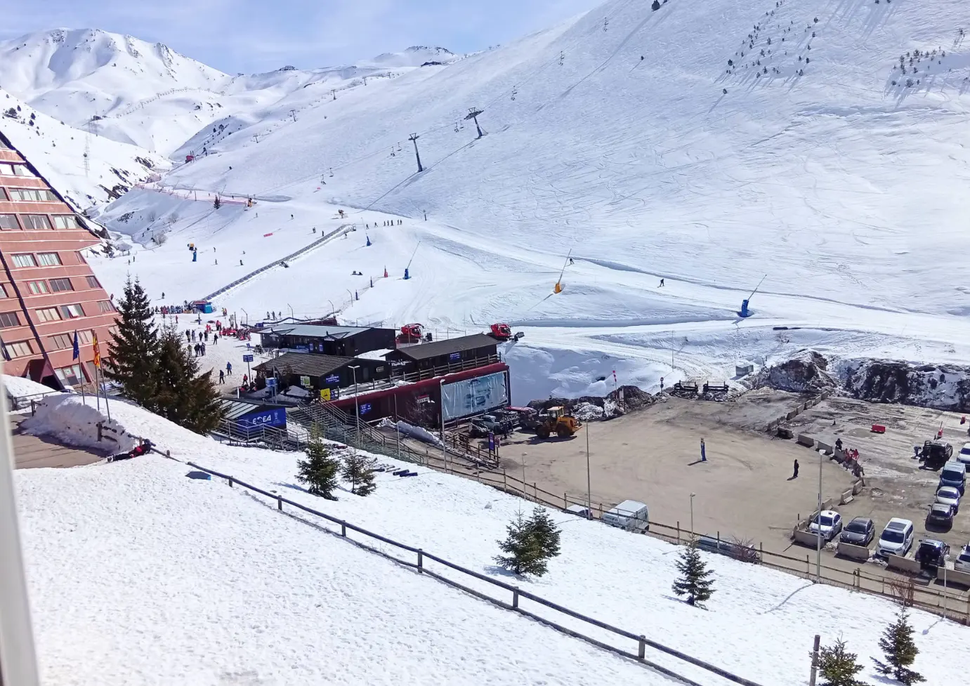 View of a snowy mountain ski resort with a large A-frame building, ski slopes, chairlifts, and a parking lot below.
