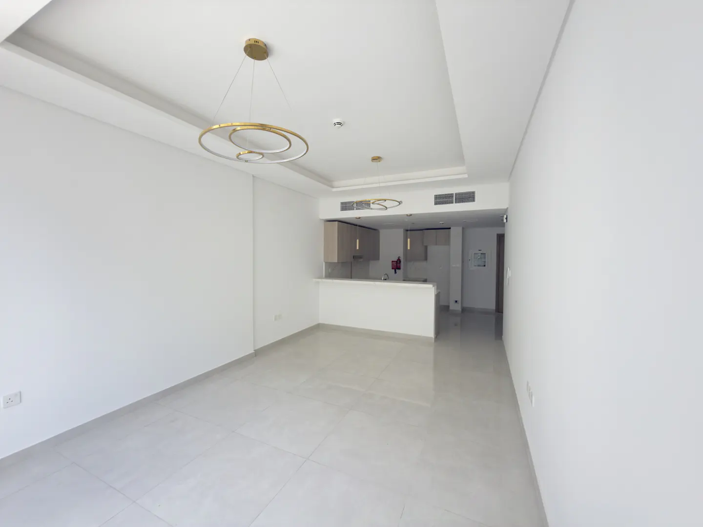 Modern open-plan living area with white walls, tiled floors, and a gold circular chandelier, leading to a small kitchen.