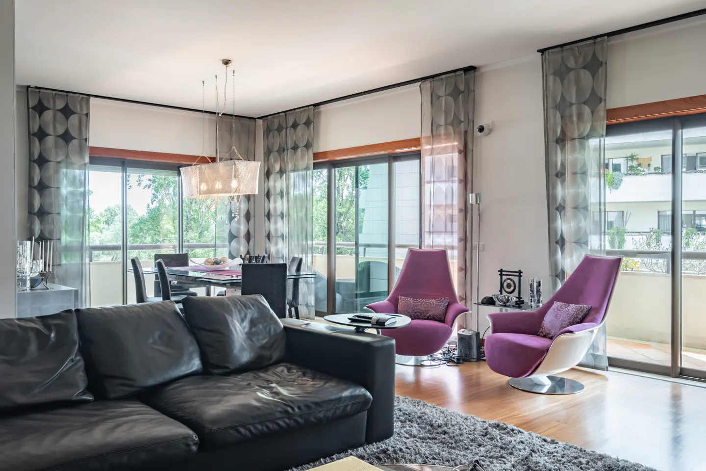 A modern living room with a black leather sofa, purple chairs, and a dining table. Large windows offer a view of greenery.