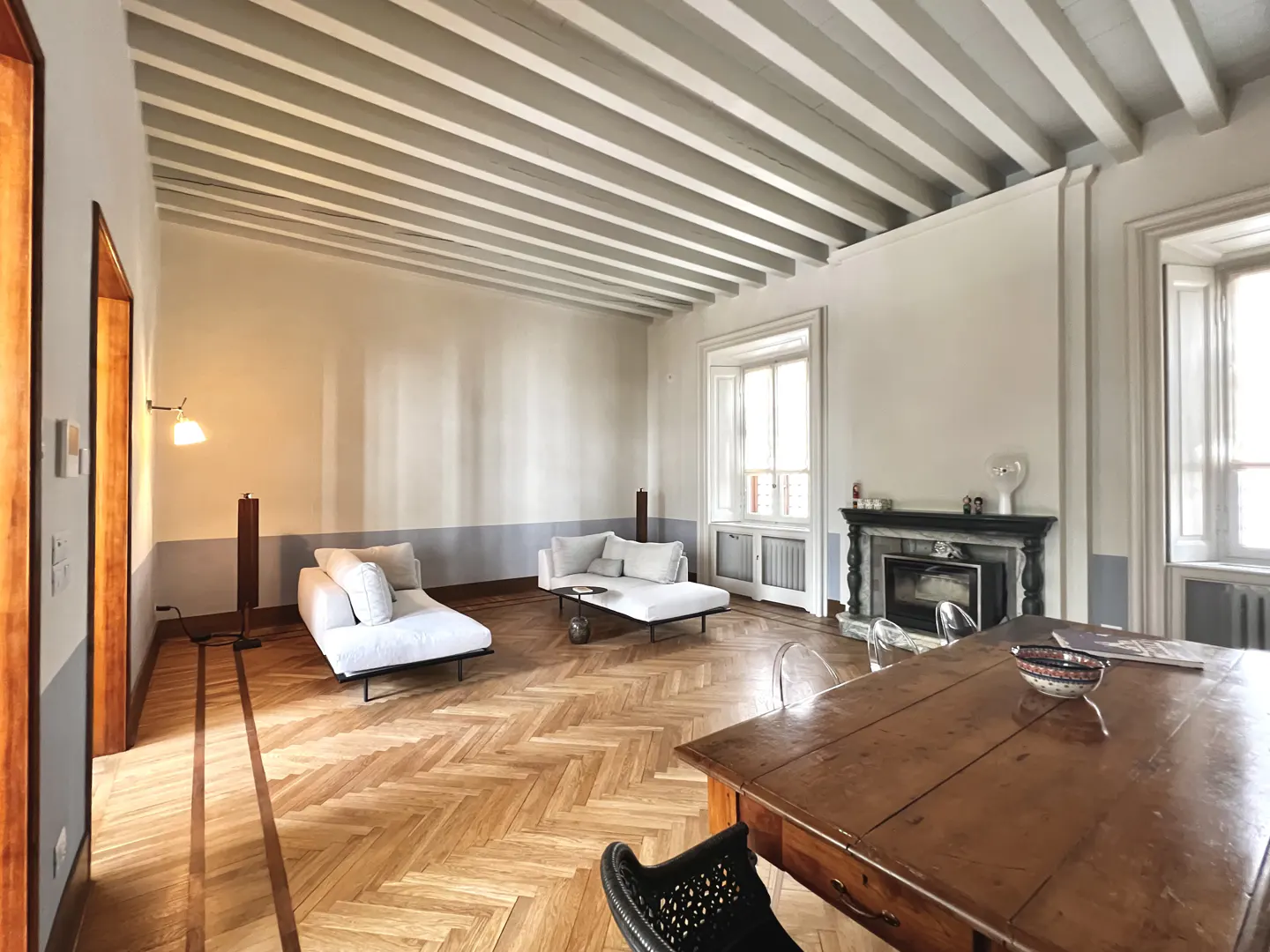 Bright living room with white walls, herringbone wood floors, and exposed beam ceiling. Two white sofas face a fireplace. A large wood table sits in the foreground.