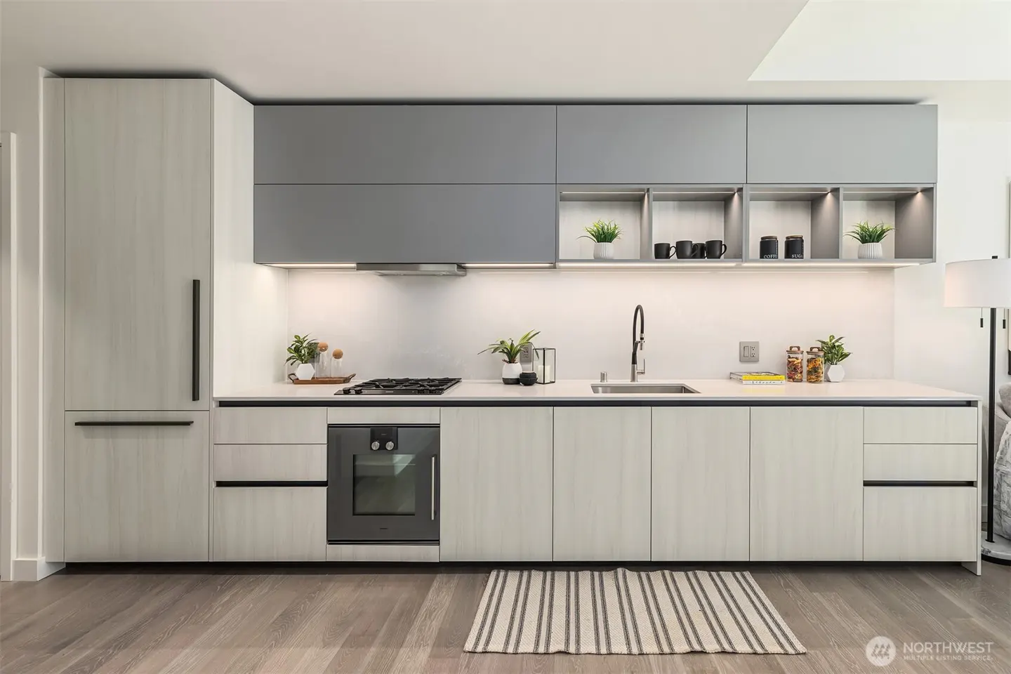 Modern kitchen with gray and light wood cabinets, white countertops, black hardware, and a striped rug on a wood floor.