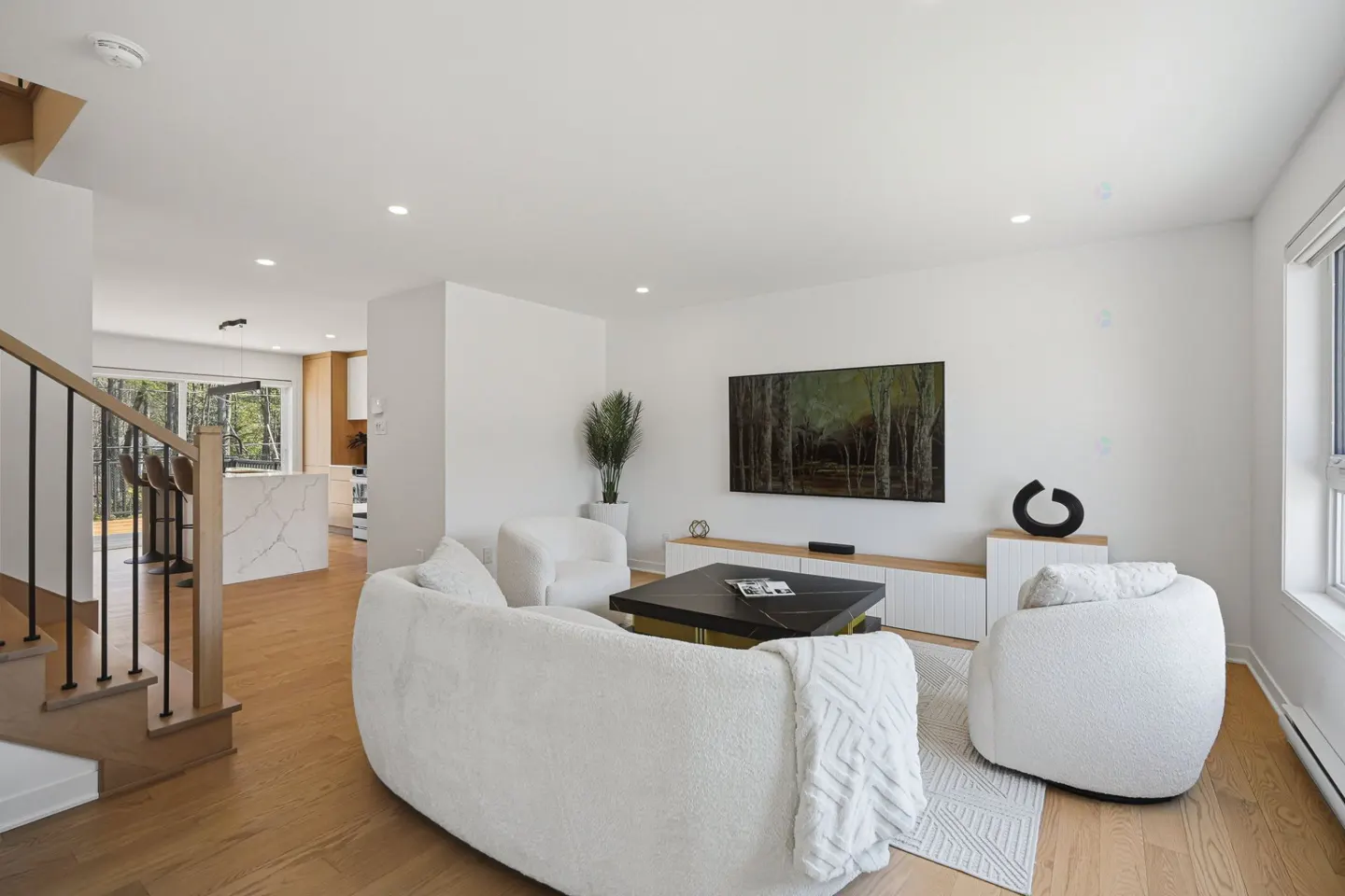 Modern living room with white curved sofas, a black coffee table, and a wall-mounted TV, overlooking a kitchen area.