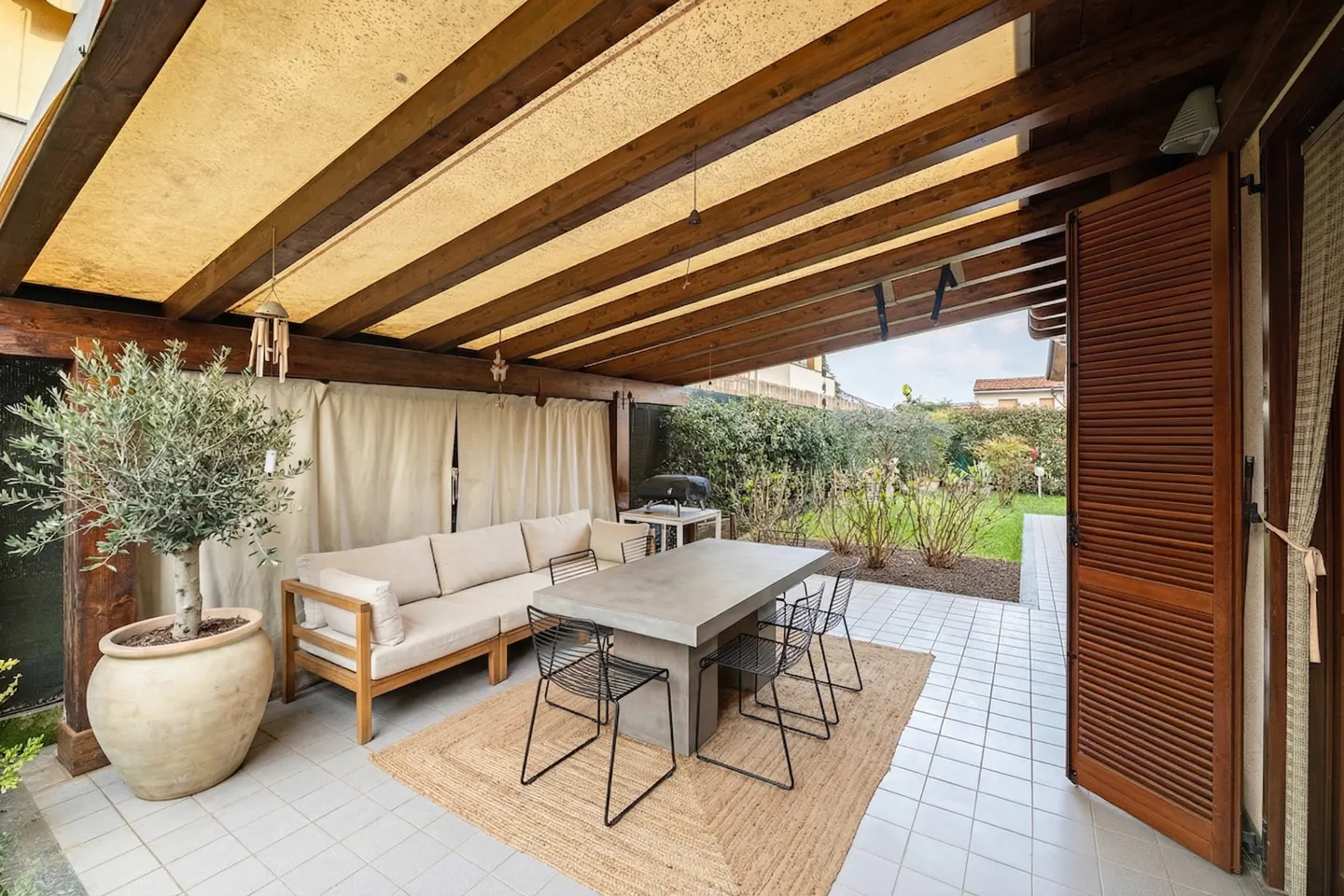 Covered patio with a beige sofa, concrete table, and black chairs on a jute rug. An olive tree sits in a pot. The garden is visible in the background.