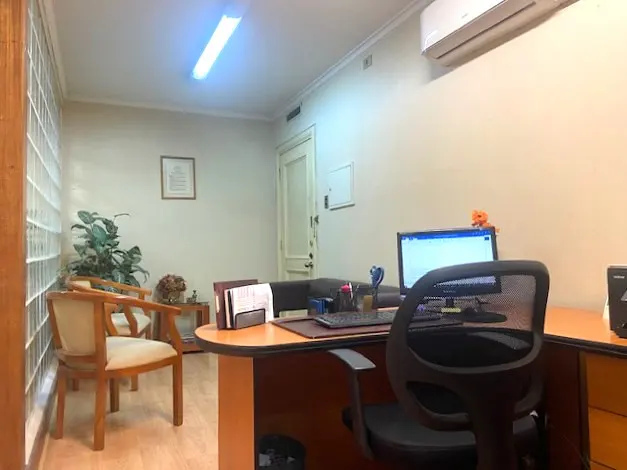 Office interior with a wooden desk, computer, and black mesh chair. Two wooden chairs and a plant are in the background.