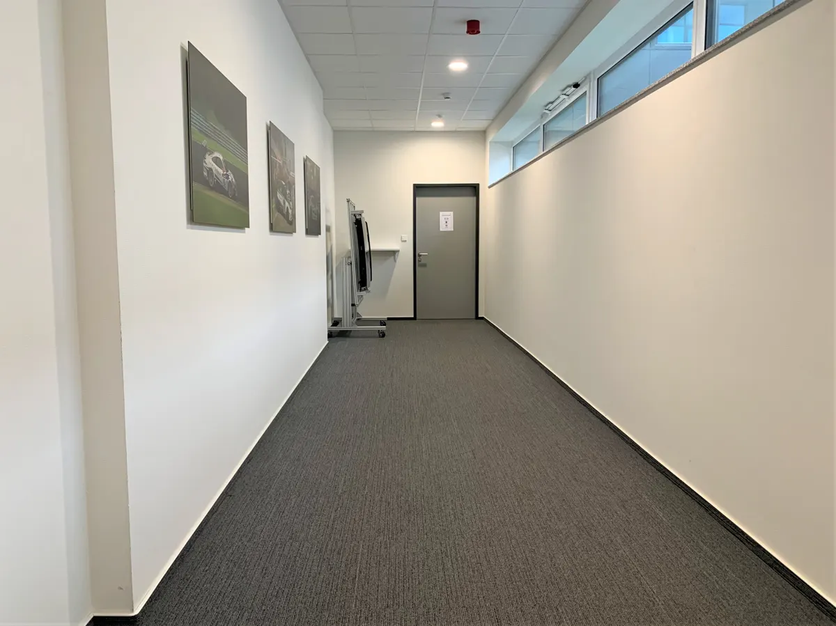A long, empty hallway with gray carpet and white walls. Pictures hang on the left wall, and a restroom door is at the end.