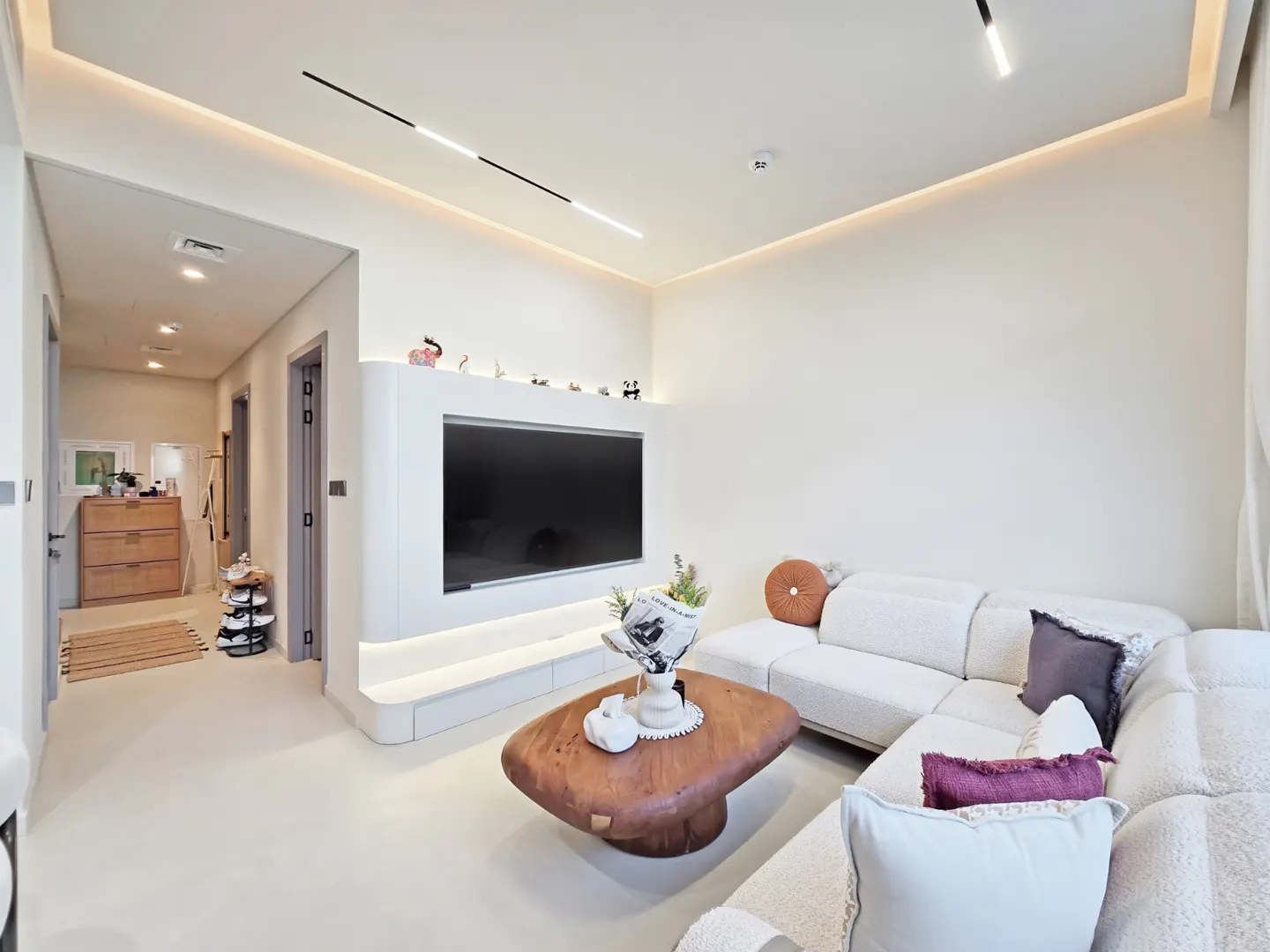 Living room with a white sectional sofa, a wooden coffee table, and a large TV on a white wall unit.