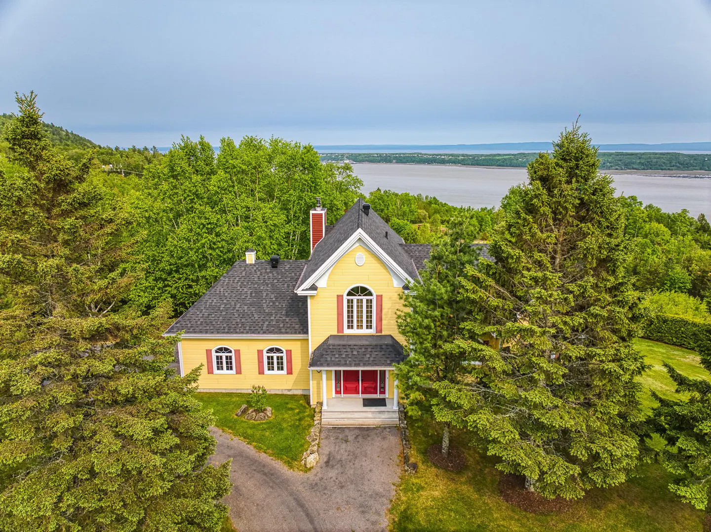 Aerial view of a yellow two-story house with a red door and trim, surrounded by green trees and a body of water in the background.