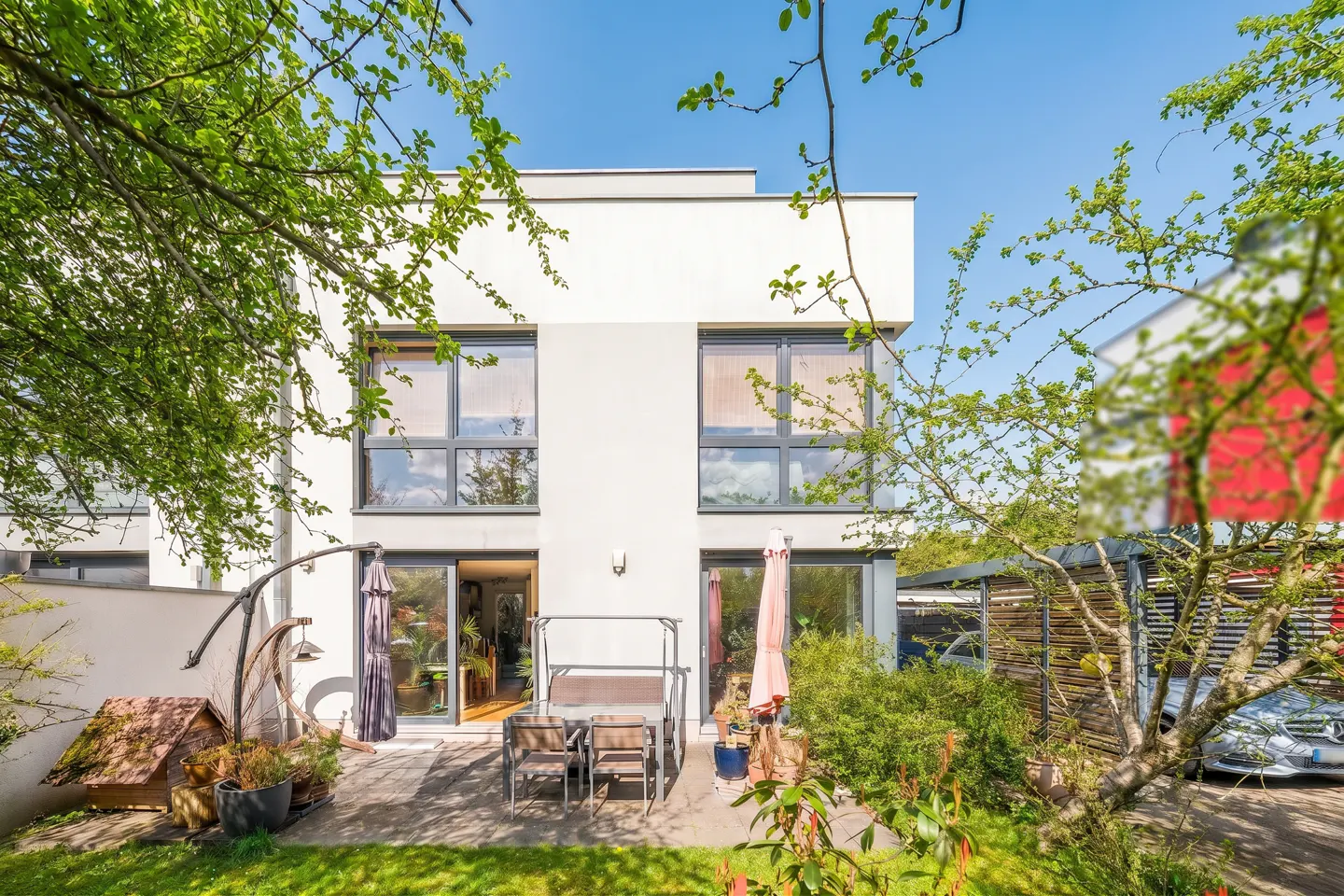 Modern two-story white house with a patio, table, chairs, and green foliage under a blue sky.
