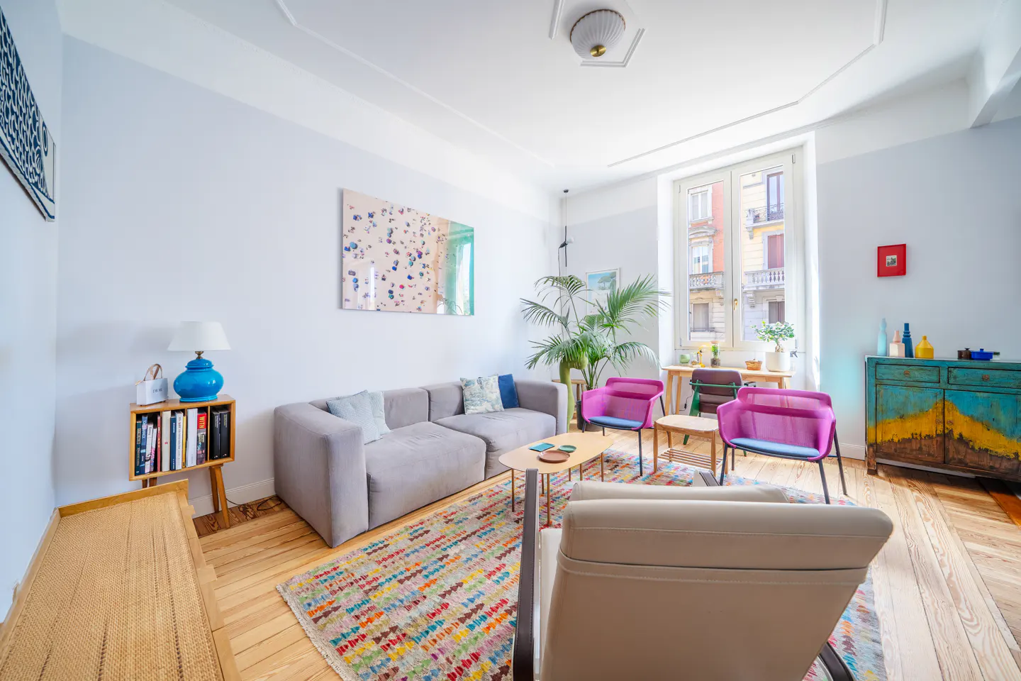 Bright living room with a gray sectional sofa, colorful rug, and pink chairs. A large window shows a building exterior.