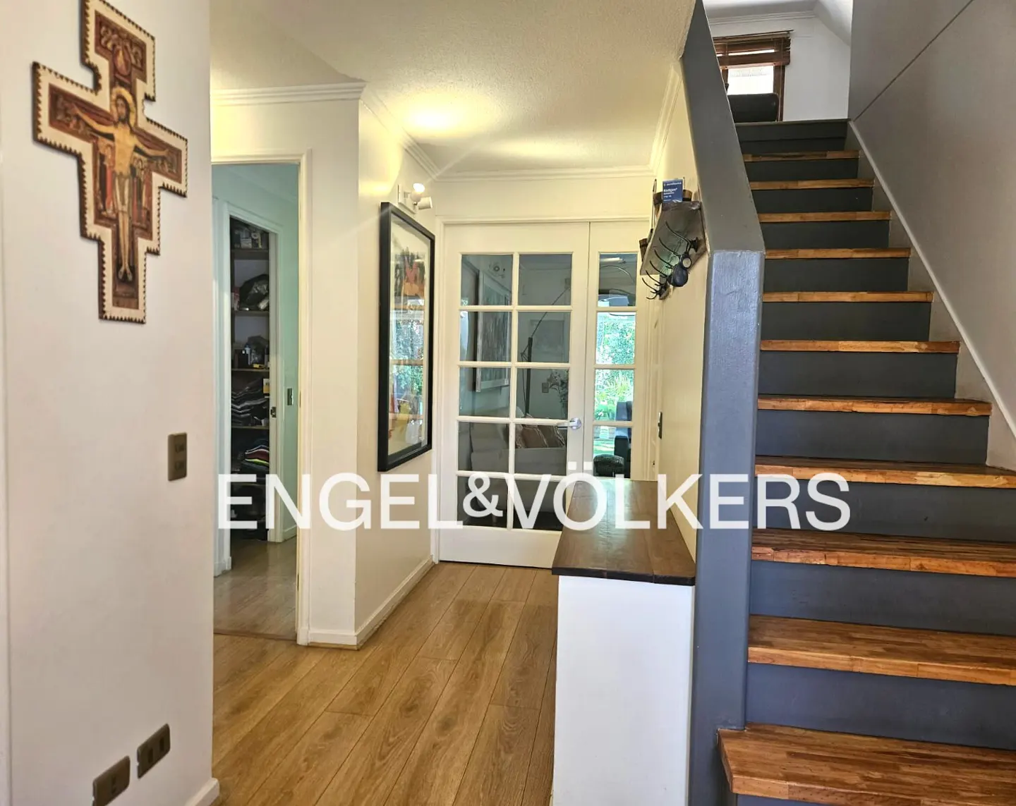 A bright hallway with wood floors, white walls, and a staircase with gray risers. A cross hangs on the wall. French doors lead to the outside.