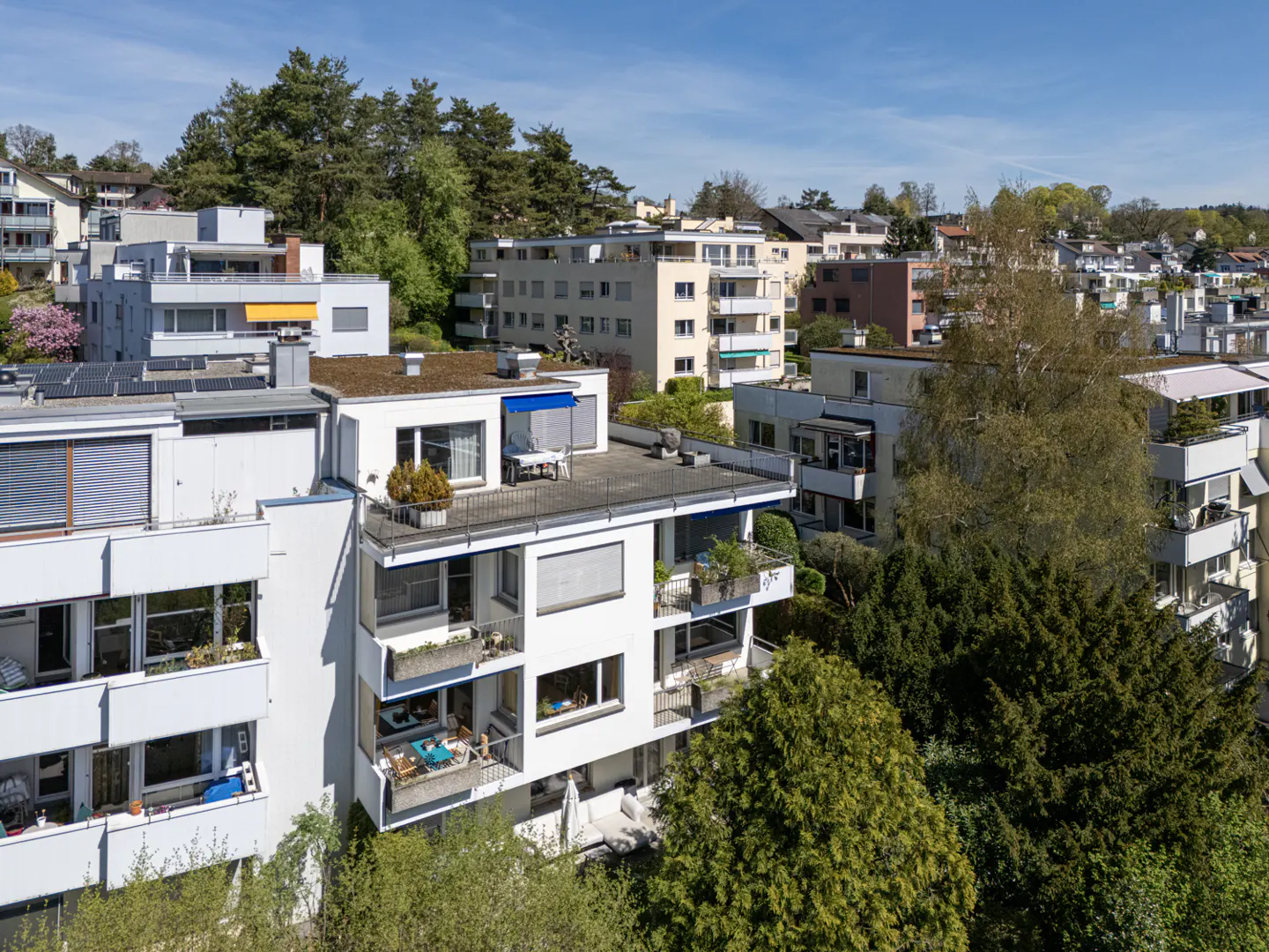 Aerial view of white apartment buildings with balconies and rooftop terraces, surrounded by green trees under a blue sky.