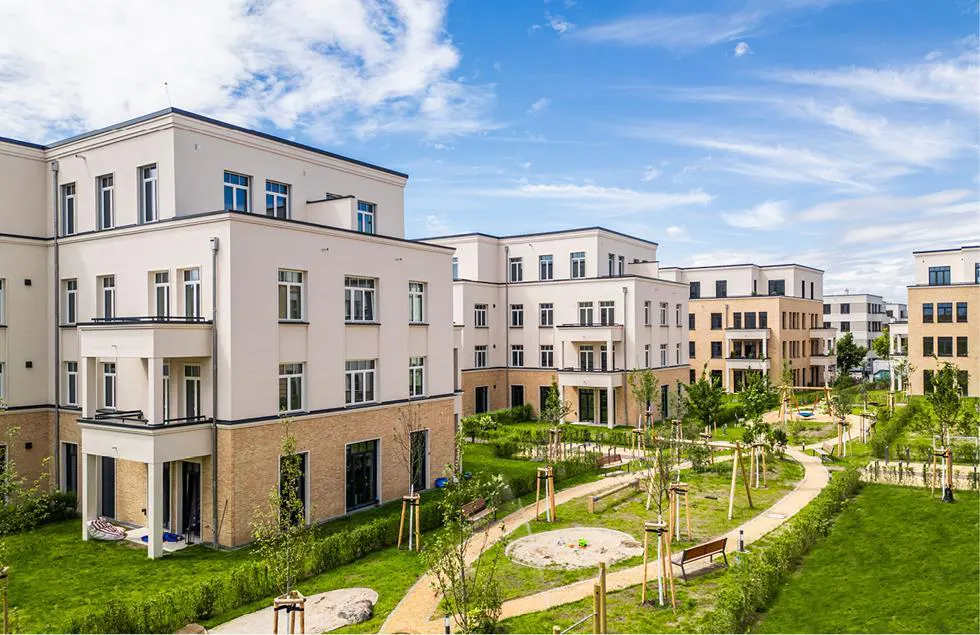 Modern apartment buildings with white facades and balconies surround a green courtyard with walking paths and trees under a blue sky.