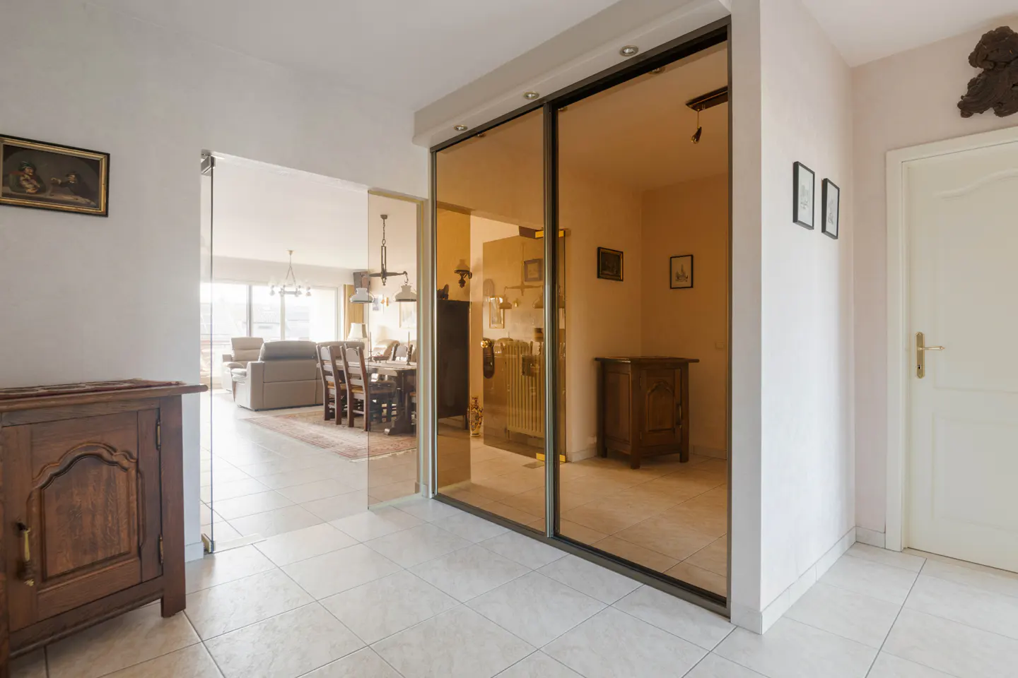 Hallway with beige tile floor, a wooden cabinet, and a view into the living room with a dining table and sofa. A mirrored sliding door reflects the interior.