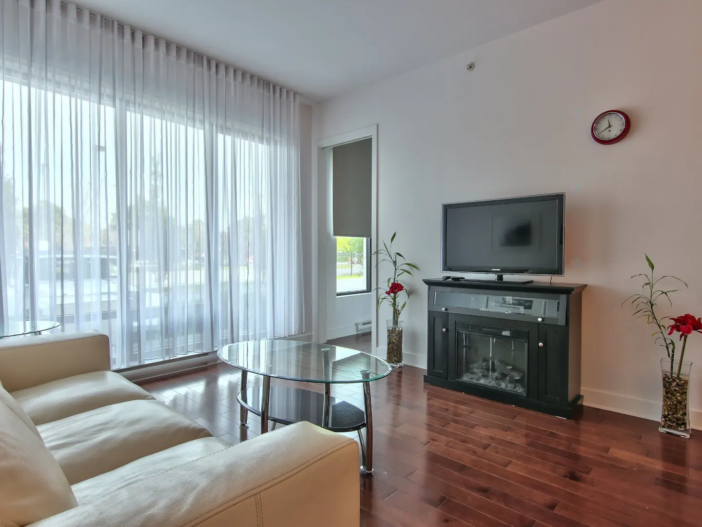 Modern living room with a white sofa, glass coffee table, TV on a black fireplace console, and floor-to-ceiling curtains.