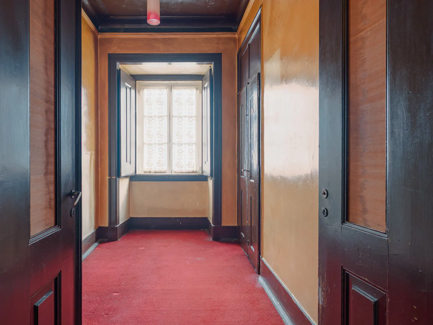 Hallway with red carpet, tan walls, and dark trim. A window with lace curtains is at the end of the hall.