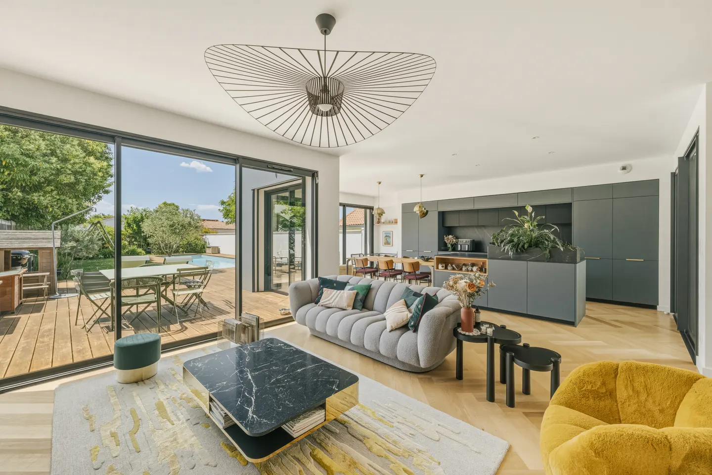 Modern open-plan living room with a grey sofa, black marble coffee table, yellow armchair, and view of a backyard pool.