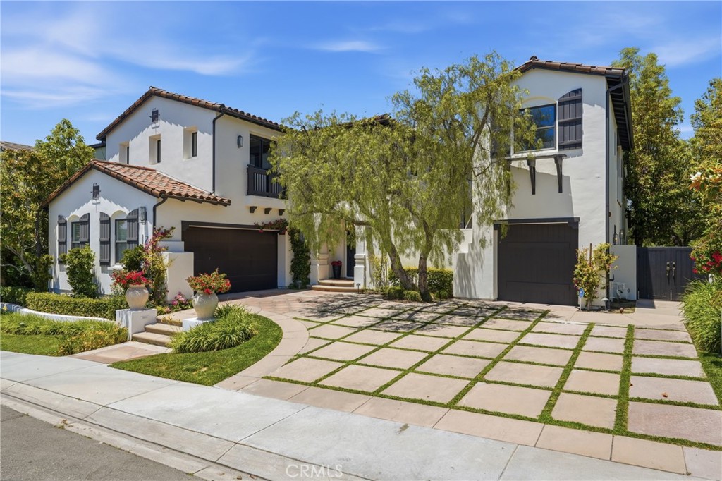 Two-story Spanish-style home with white stucco, dark garage doors, and a grid-patterned driveway under a blue sky.