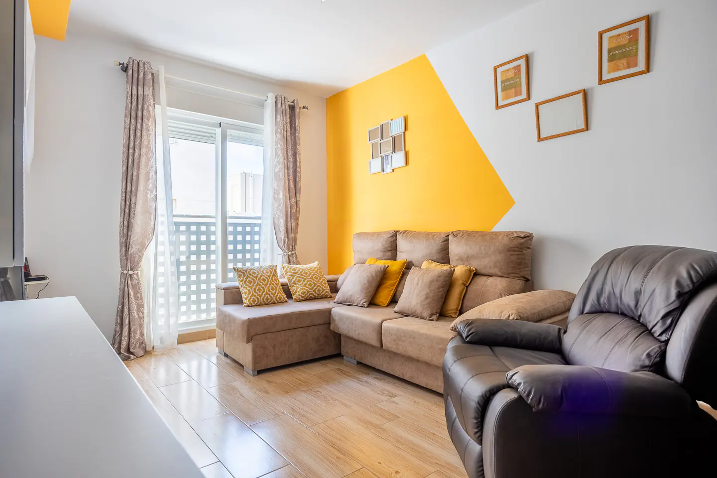 Living room with brown sectional sofa, leather recliner, and yellow accent wall. Pictures hang above the sofa. Curtains frame a window.