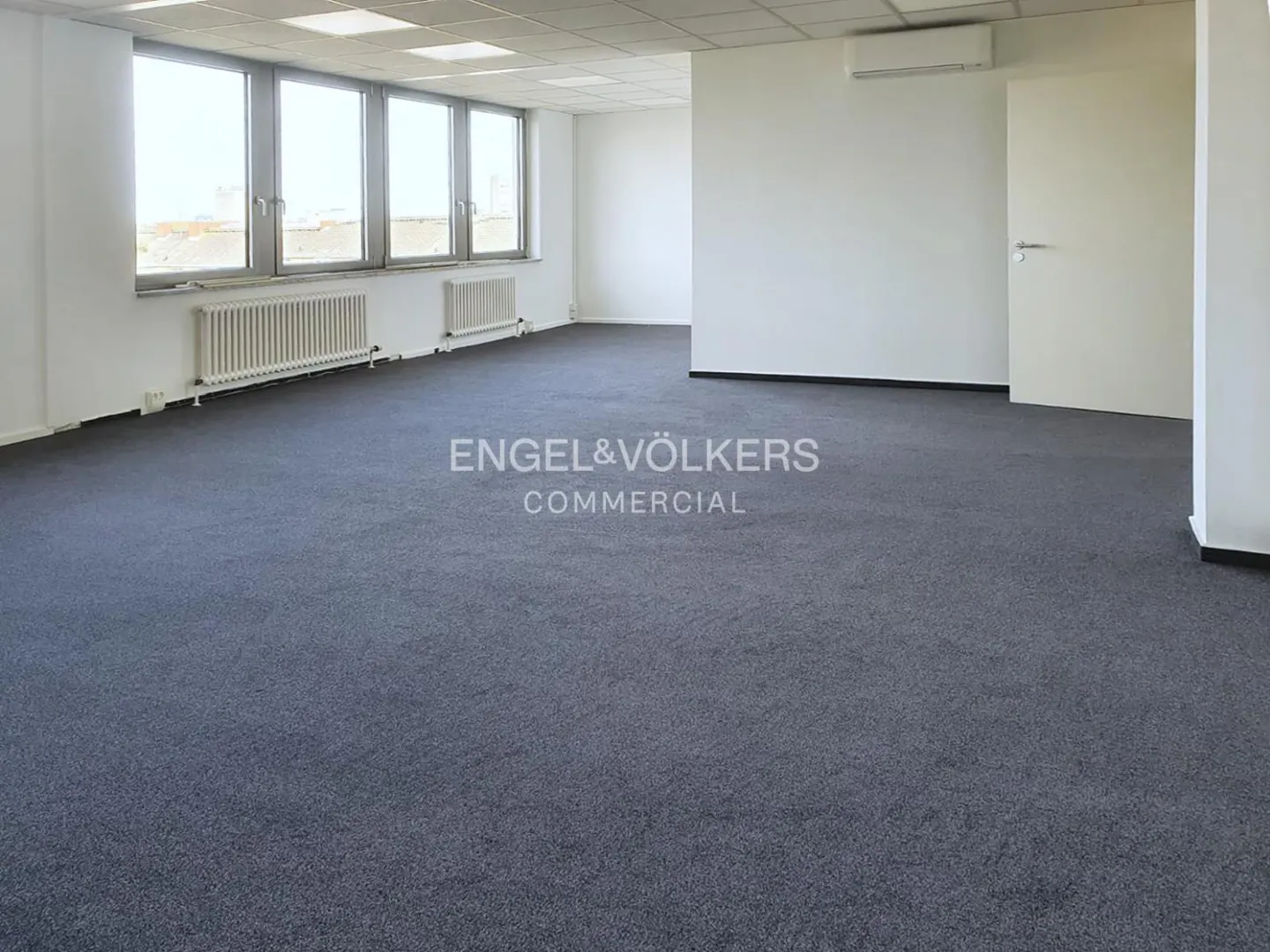 Empty office space with gray carpet, white walls, and large windows. Radiators sit beneath the windows. A white door is on the right.
