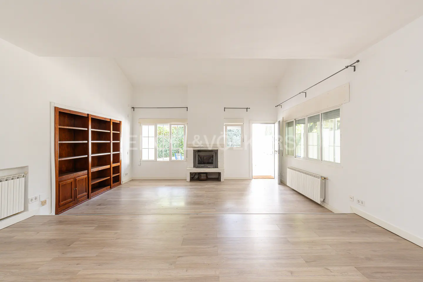 Bright, empty living room with light wood floors, white walls, a fireplace, and a large, dark wood bookcase.