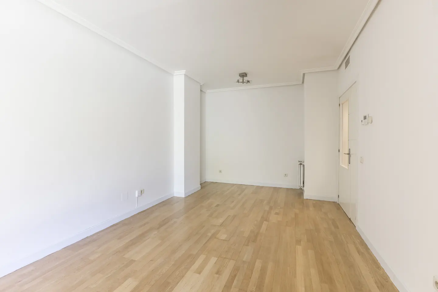 Bright, empty room with light wood floors and white walls. A door is visible on the right.