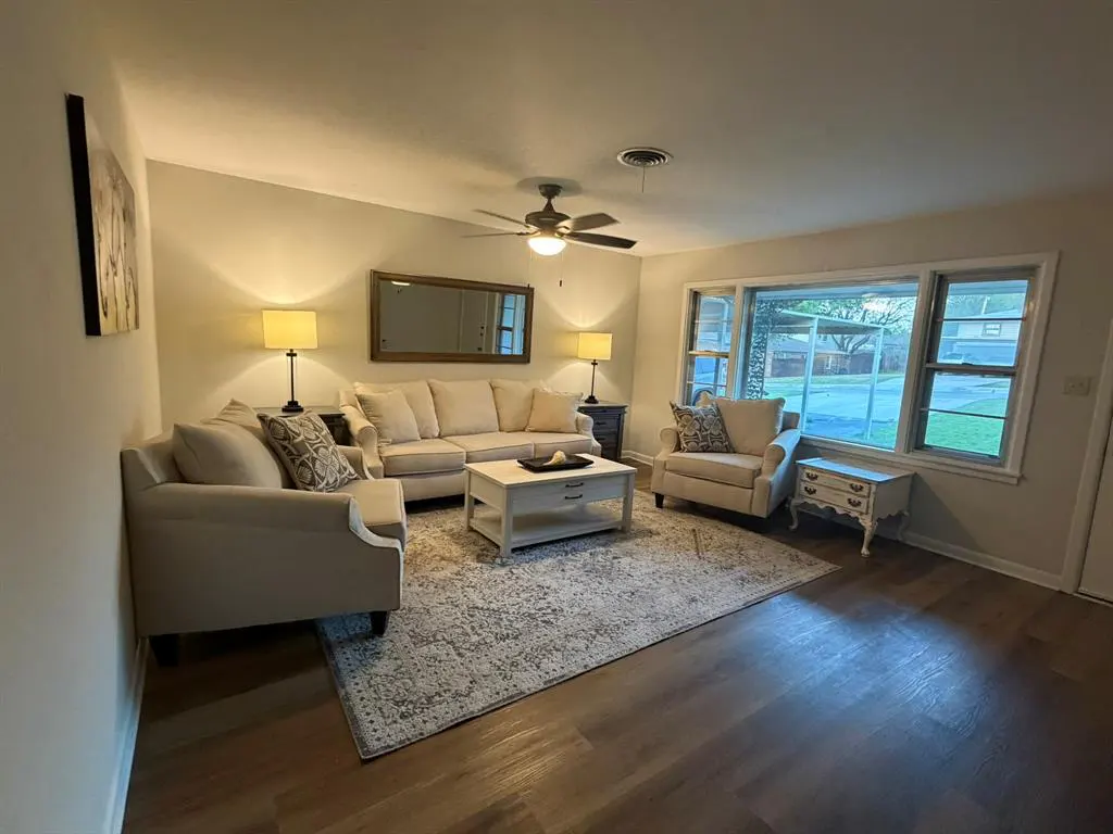 Living room with beige sofa, two armchairs, and a white coffee table on a patterned rug. A ceiling fan and large window provide light.