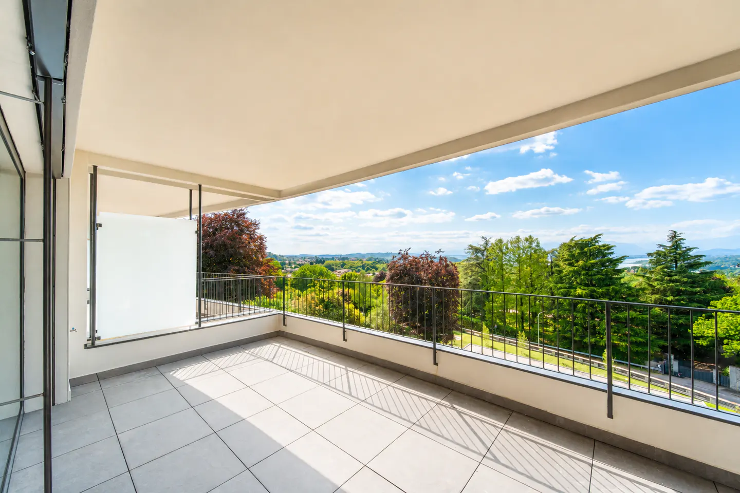 A bright, airy balcony with gray tile flooring overlooks a lush green landscape under a blue sky.