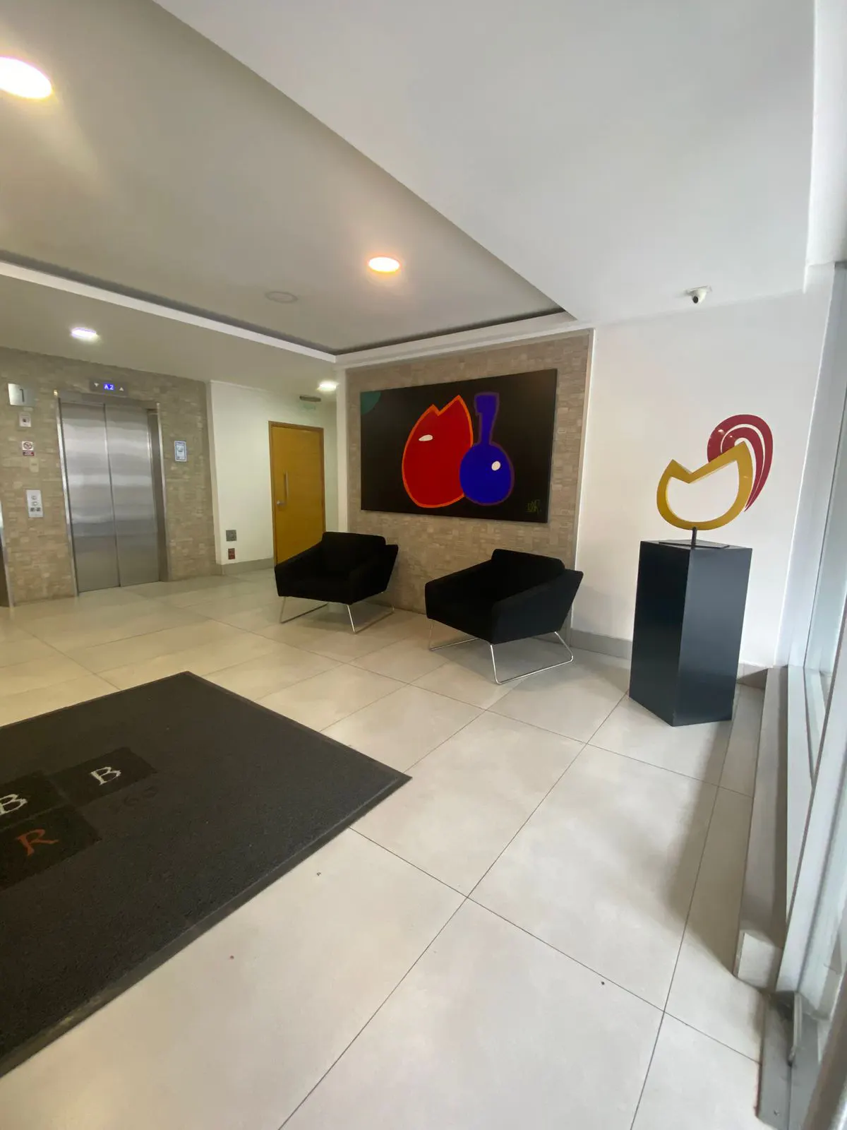 Lobby area with two black lounge chairs, abstract art, and a sculpture on a black pedestal. An elevator and a door are visible in the background.
