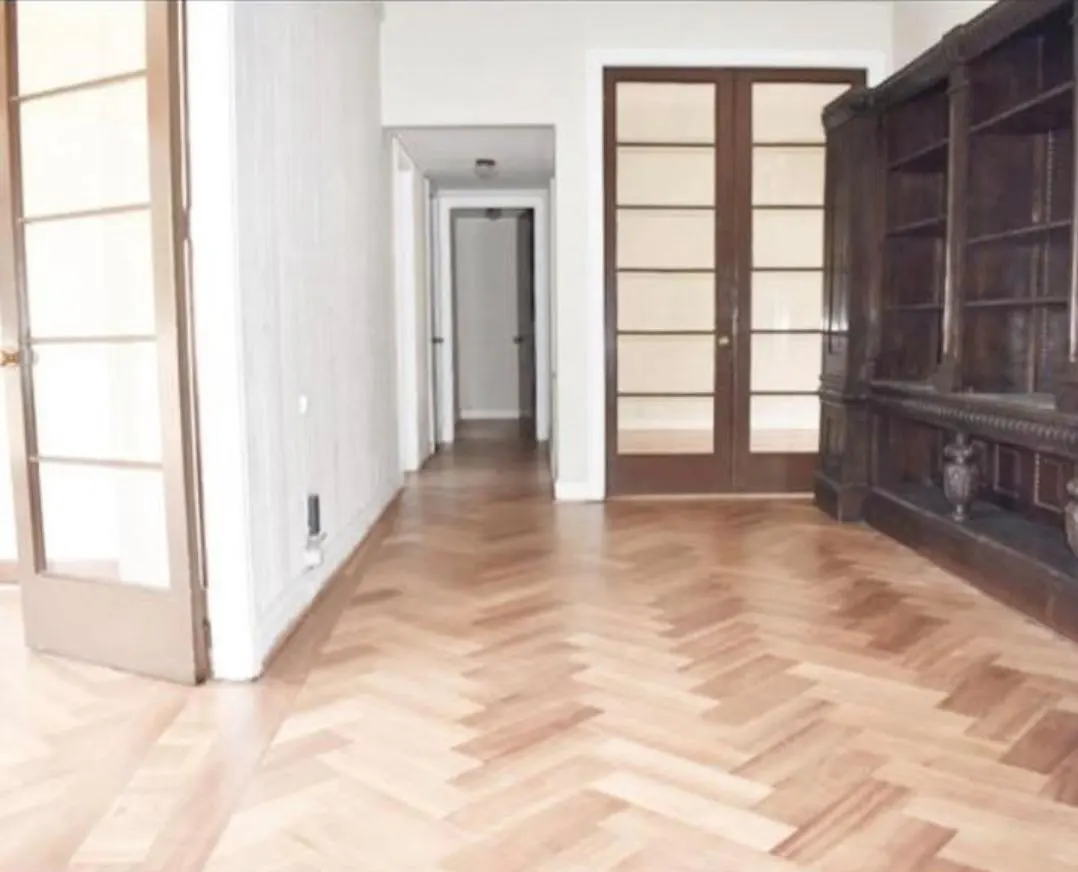 An interior view of a room with herringbone wood floors, white walls, a dark wood bookcase, and glass paneled doors.