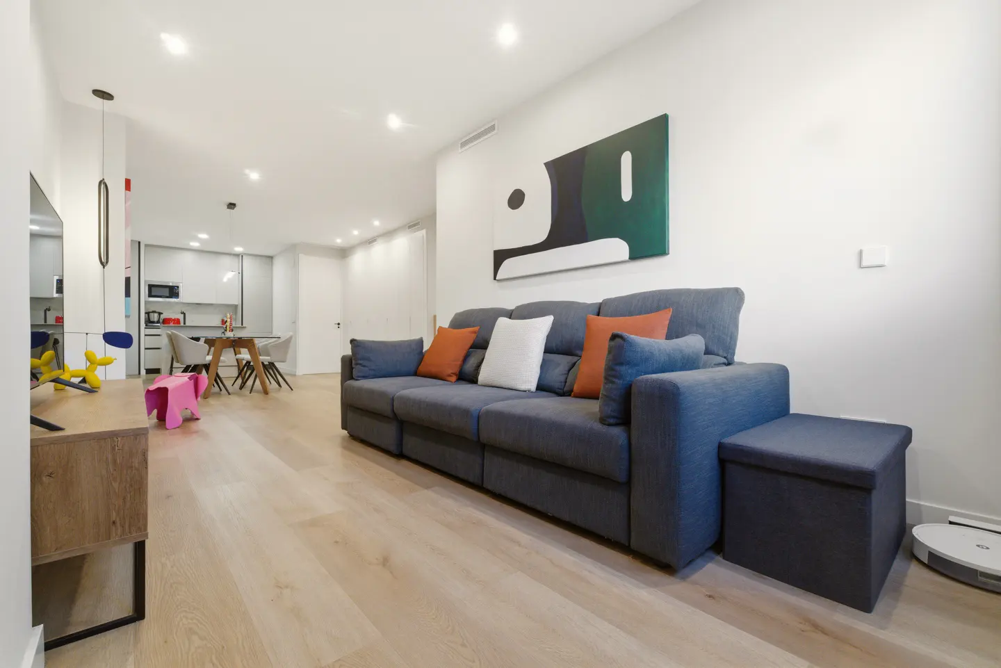 Bright living room with a blue sofa, orange pillows, and abstract art. A kitchen and dining area are visible in the background.